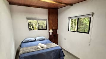 Een slaapkamer met een bed, twee ramen, een houten plafond en een kamerkleed met een patroon.
