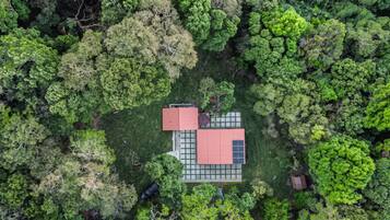 An aerial view of a house with a red roof, surrounded by dense green forest.