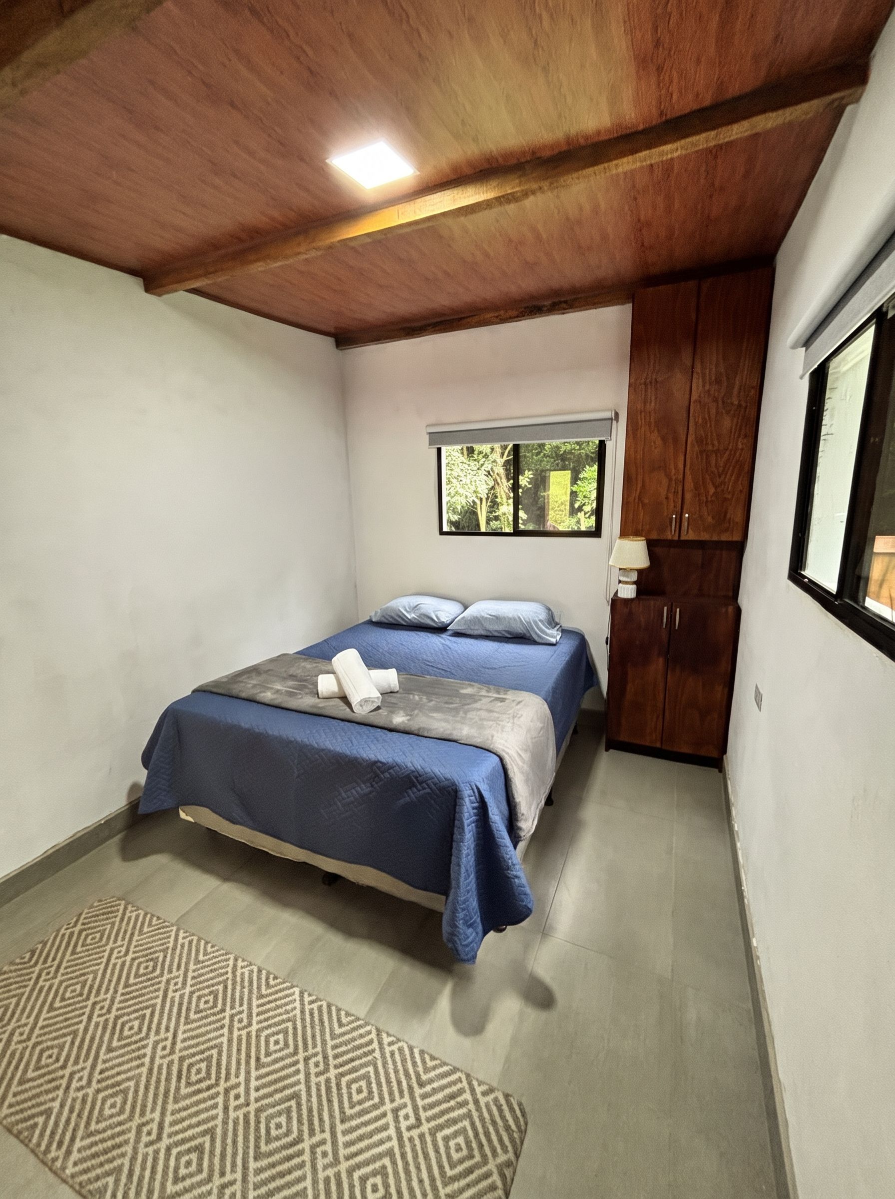 A bedroom with a bed, a window, a wooden ceiling, and a patterned rug.