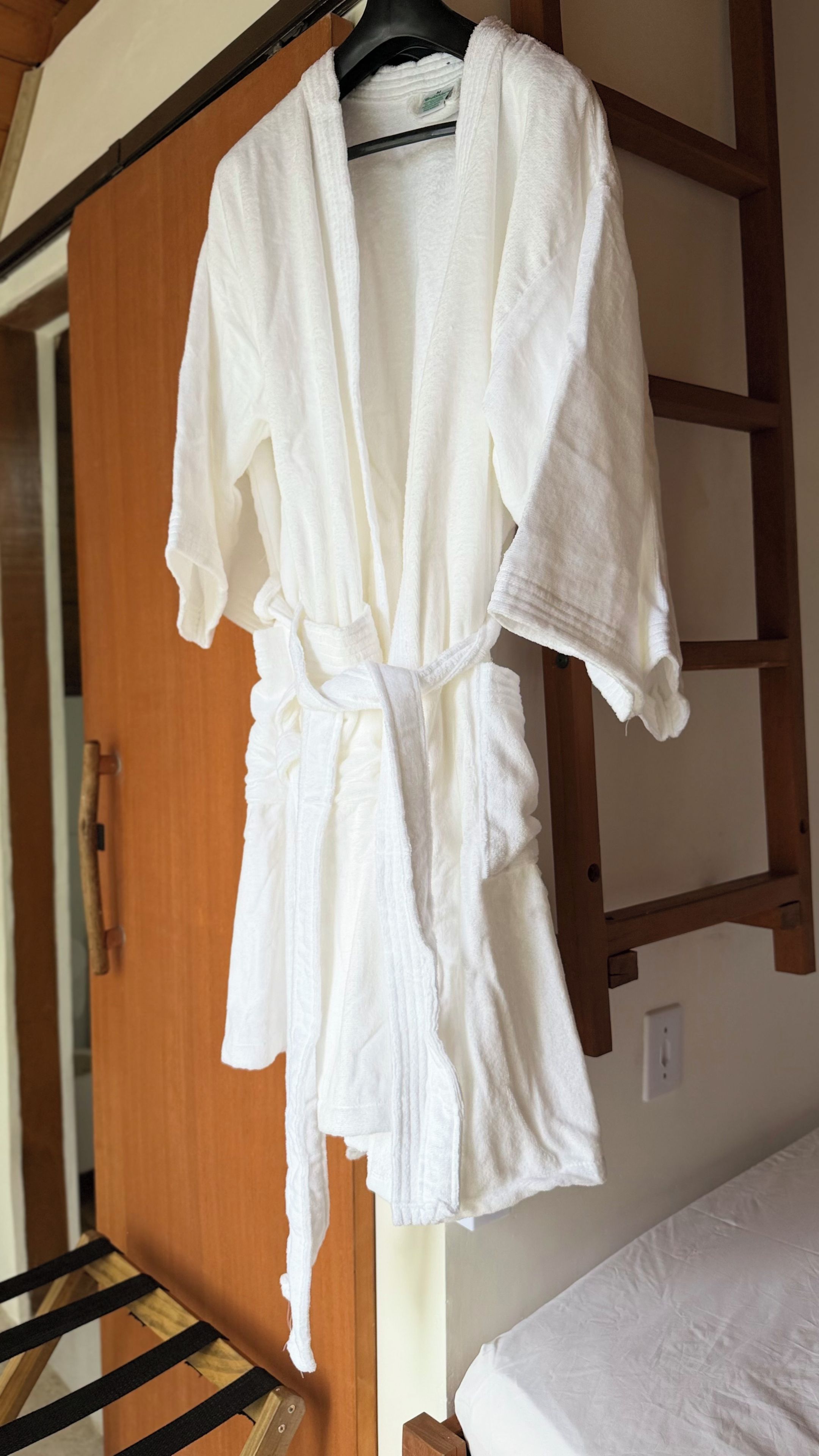 Un albornoz blanco colgado en una percha de madera, en una habitación con un armario de madera y una cama con colchón blanco.