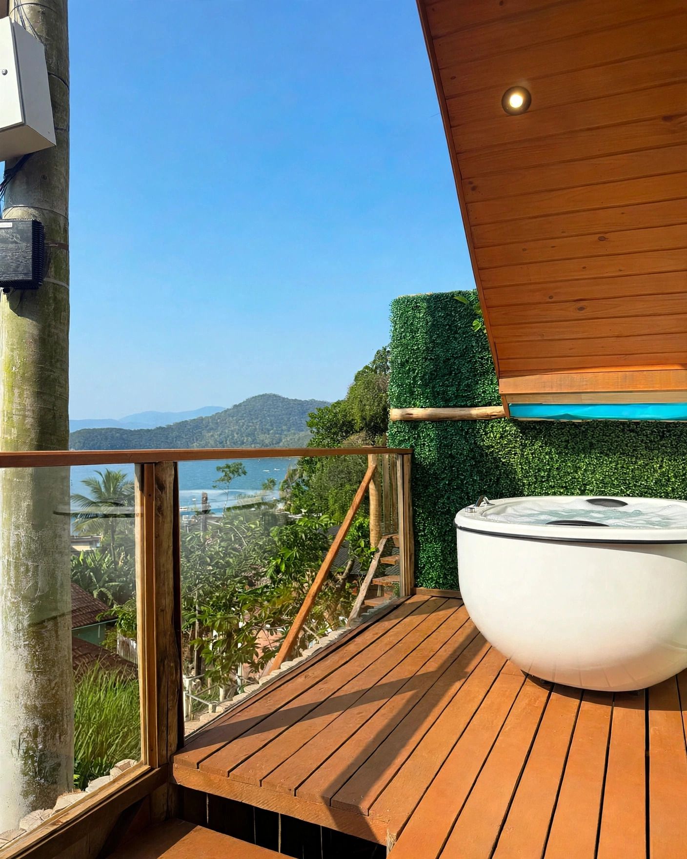 Eine Holzterrasse mit Whirlpool, Glastrennwand und Blick auf eine Küstenlandschaft.