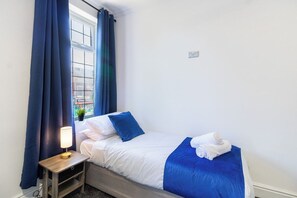 A single bed with white and blue bedding, a bedside table with a lamp, and a window with blue curtains. - Spacious Central House | Parking | Old Trafford (Stretford)