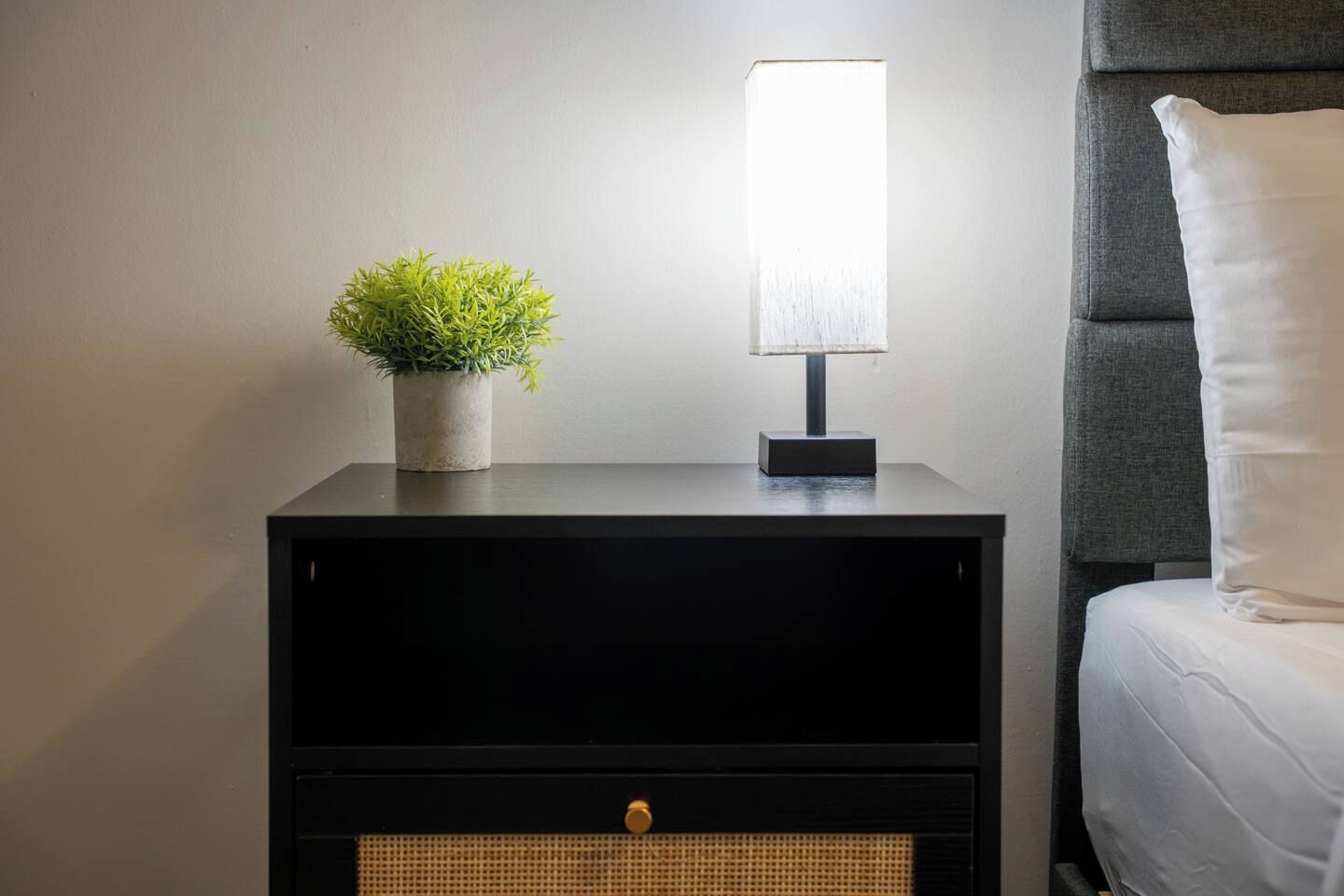 A black bedside table with a lamp, a potted plant, and a drawer.