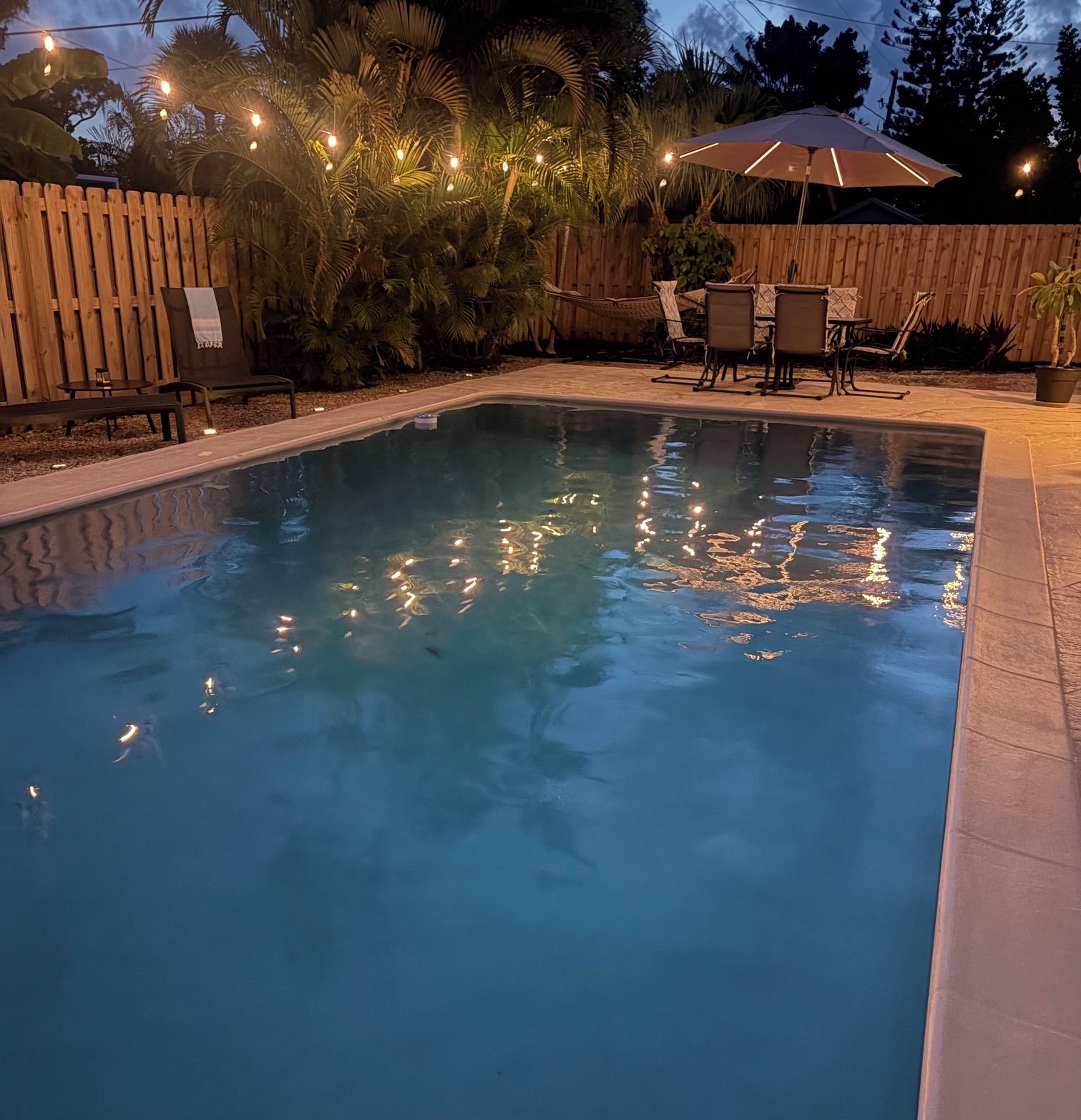 A swimming pool with a wooden deck, surrounded by palm trees and a wooden fence.