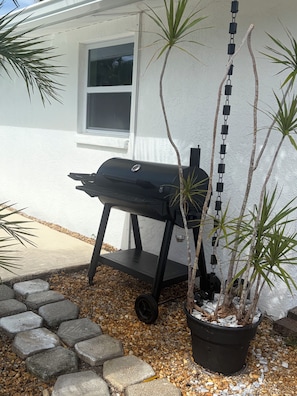 A barbecue grill with a chain hanging from it, placed on a gravel area with stepping stones leading to a house with a window. - Relax, Swim & Explore! Cozy Venice Home near the beach. (Venice)
