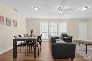Ein modernes Wohnzimmer mit Essbereich, Deckenventilator und großen Fenstern mit Jalousien.