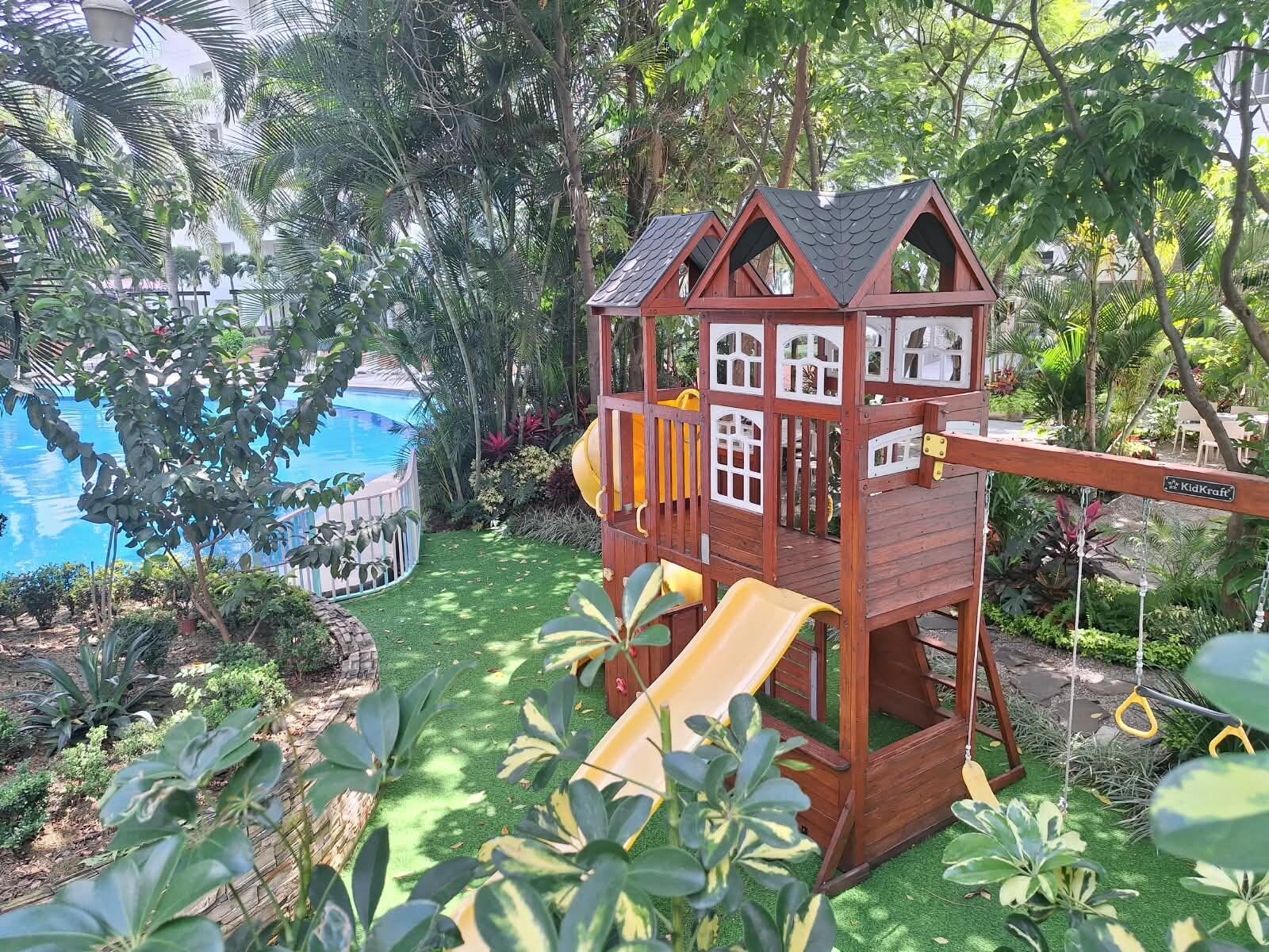 A wooden playhouse with a slide, surrounded by lush greenery and a pool.