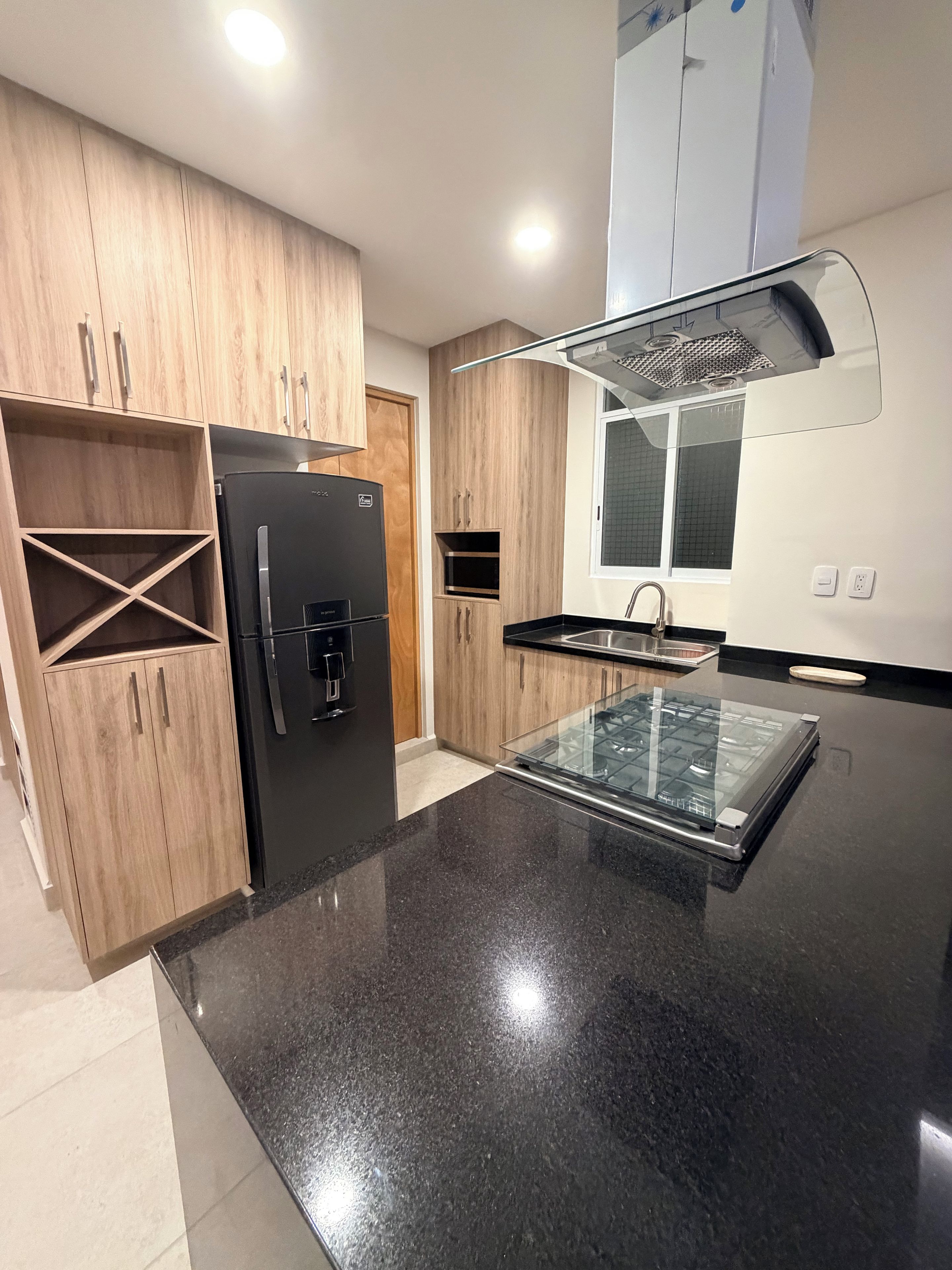 A modern kitchen with wooden cabinets, a black refrigerator, a glass-top island, and a window above the sink.