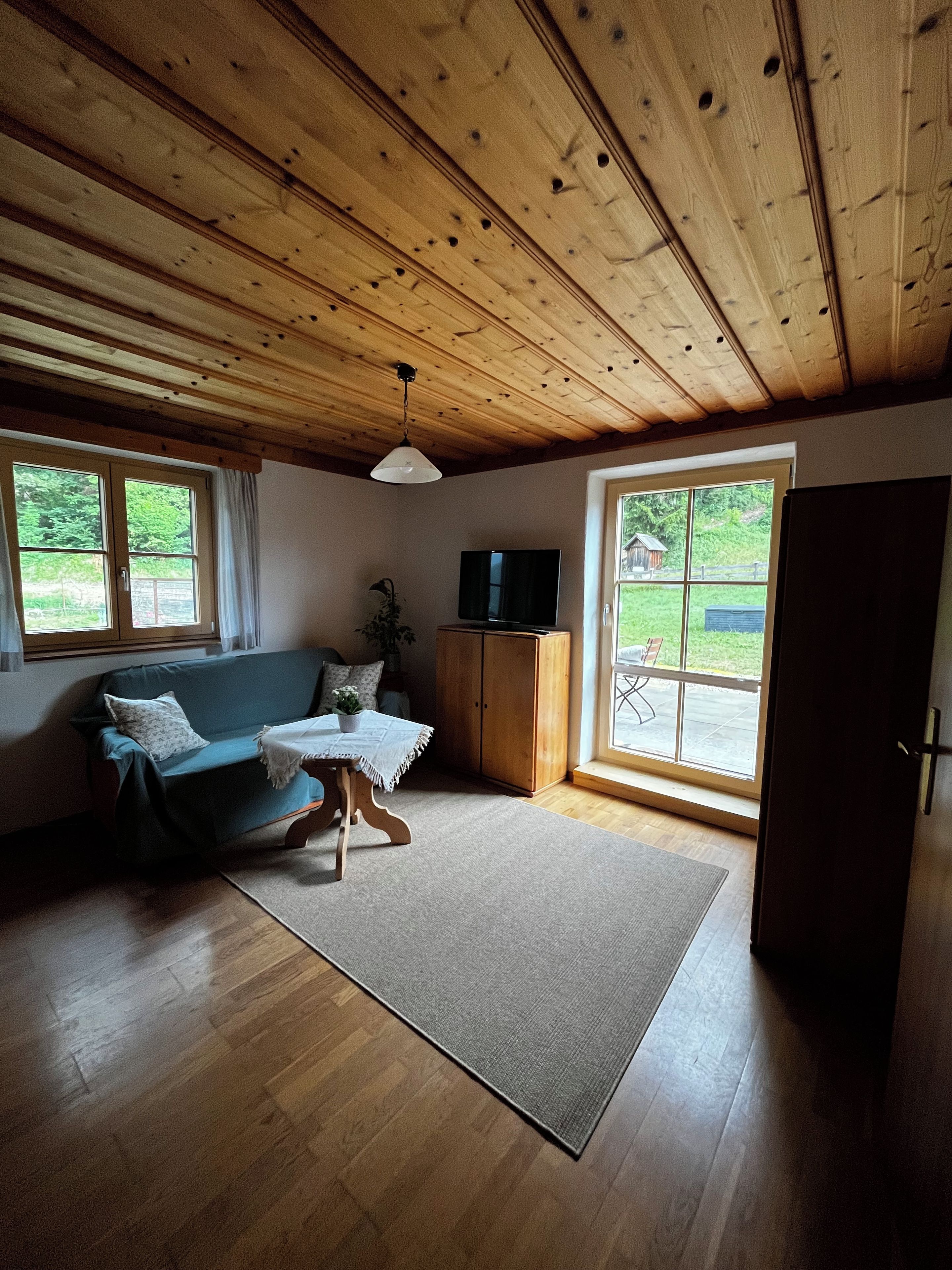 Un salon chaleureux avec un plafond en bois, un canapé, un téléviseur et une vue sur l’extérieur grâce à de grandes fenêtres.