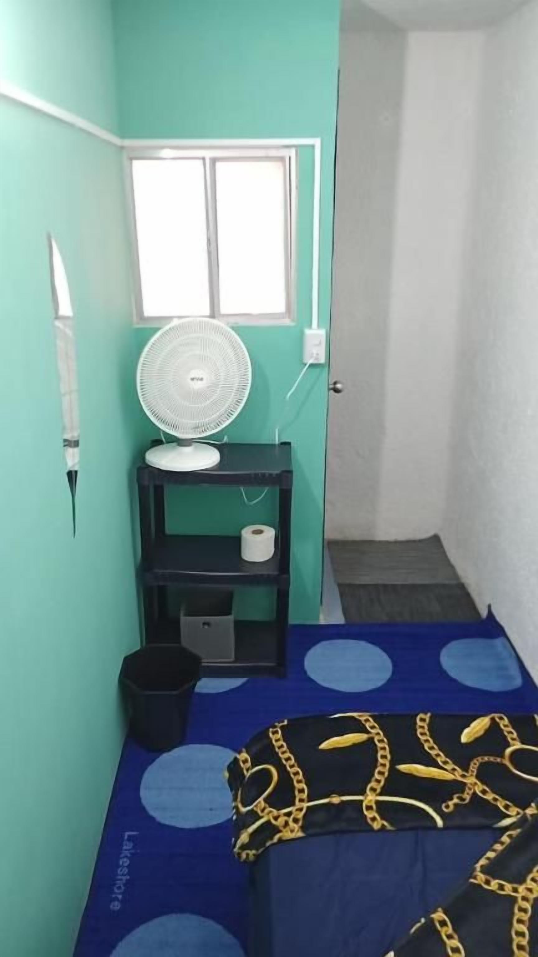 A small room with a bed, a fan on a shelf, a window, and a door leading to another room.