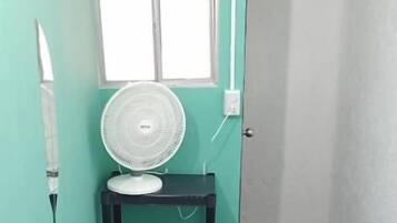 A small room with a bed, a fan on a shelf, a window, and a door leading to another room.