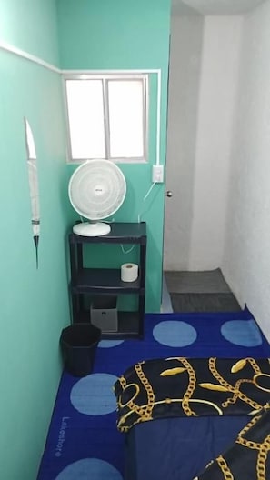 A small room with a bed, a fan on a shelf, a window, and a door leading to another room. - Rooms del Bosque Studios (MORELIA)