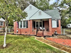 A single-story house with a brick exterior, a front porch with outdoor seating, and a flagpole with an American flag. - Cozy & Luxurious Whole Home Near Ft. Benning and Uptown (Columbus)