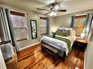 Quarto com cama, mesa de cabeceira, rodapé, piso de madeira, ventoinha no teto e janelas com persianas.