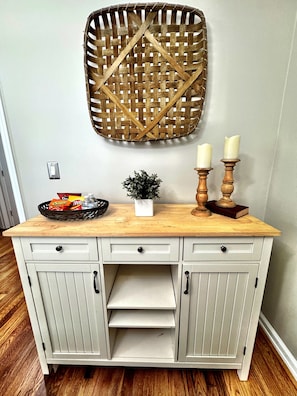A white wooden cabinet with drawers and shelves, a woven basket on top, and two candles on a wooden stand. - Cozy & Luxurious Whole Home Near Ft. Benning and Uptown (Columbus)