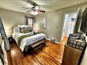 Ein Schlafzimmer mit einem Bett, einem Deckenventilator, einem Nachttisch, einer Lampe, einem Mülleimer und einem Fernseher.