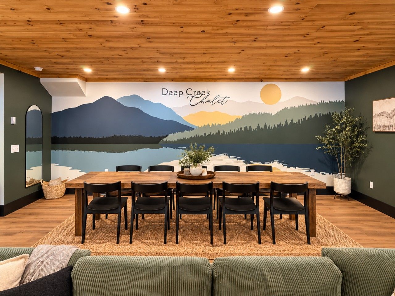 A dining area with a wooden table and chairs, a mural of a mountain landscape, and a plant on the wall.
