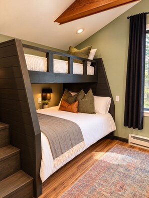 Una cama litera con cabecera, una mesita de noche con lámpara, una alfombra estampada y una ventana con cortinas.