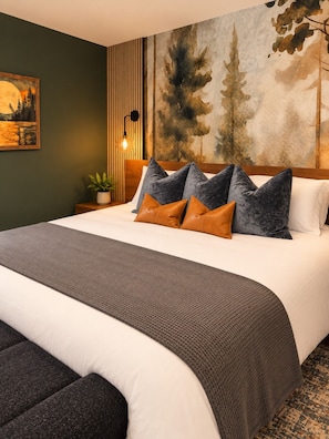 Una cama bien tendida con un edredón gris texturizado, un cabecero con una lámpara vertical y un cuadro de un paisaje forestal en la pared.