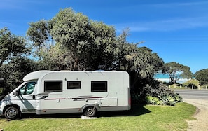 Una caravana estacionada en un área verde, con árboles y un cielo despejado al fondo.