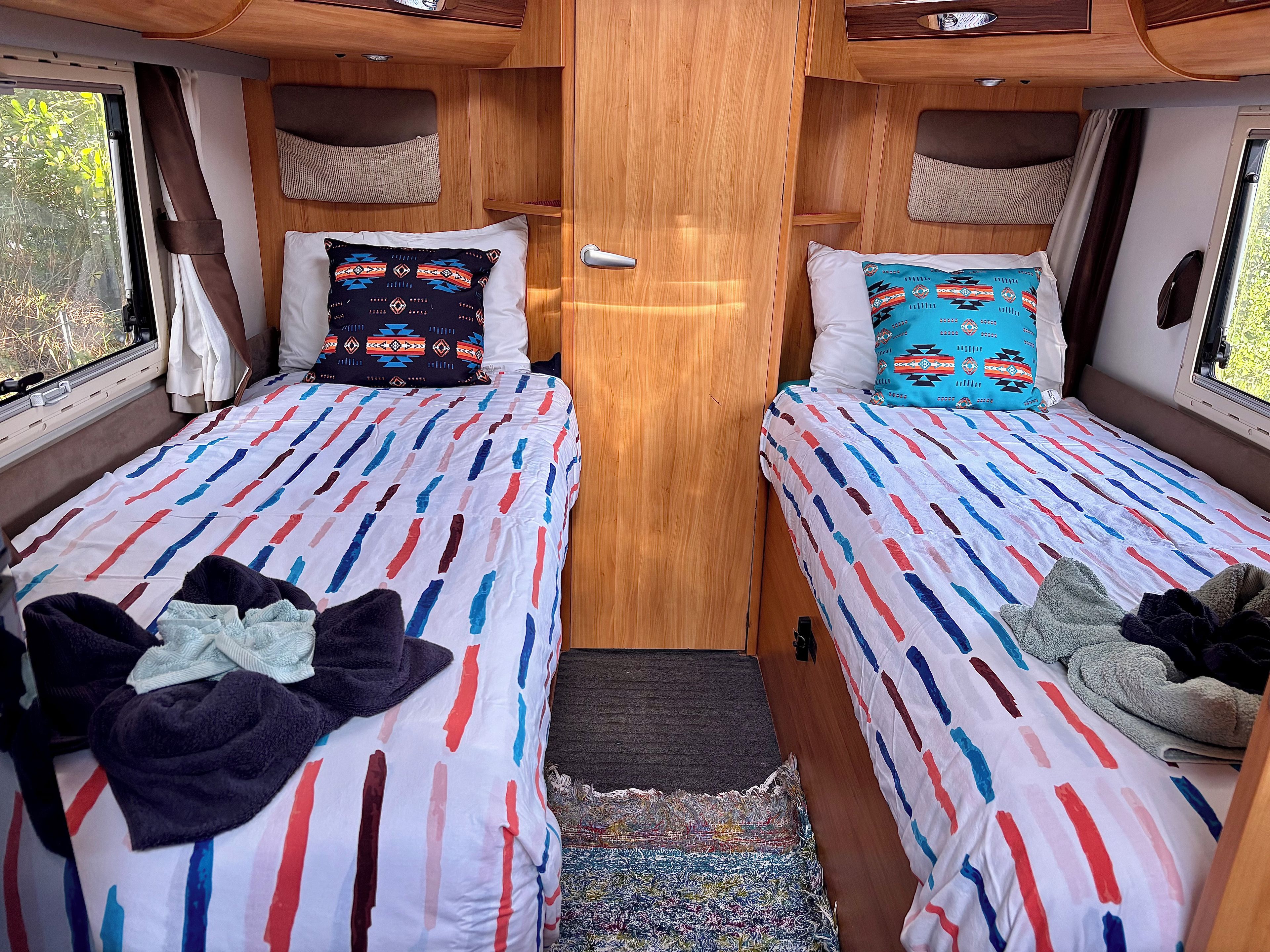 Un intérieur de camping-car compact comprenant deux lits, chacun agrémenté d’une couvre-lit à motifs et de coussins, une porte centrale et une fenêtre sur un des côtés.
