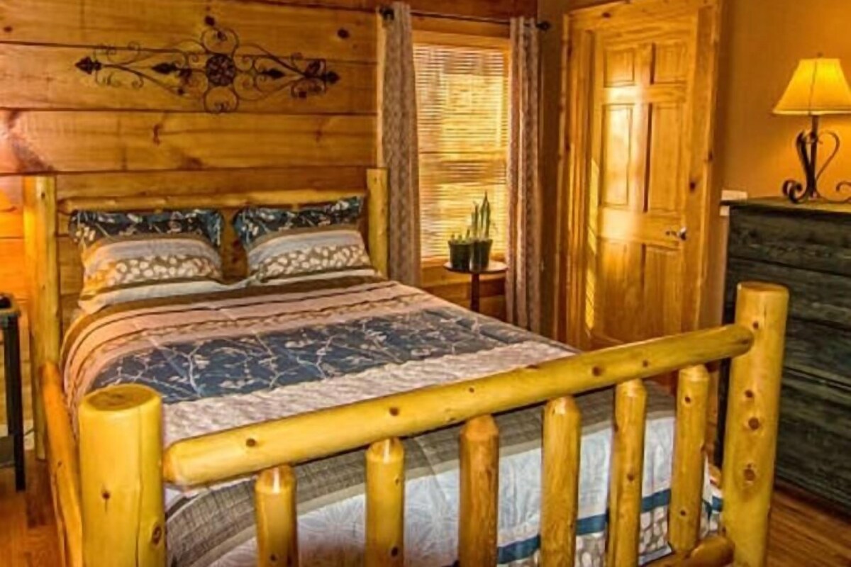 A wooden cabin interior with a bed, a nightstand, a lamp, and a door.