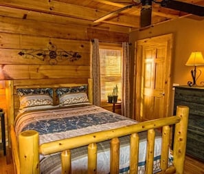 A wooden cabin interior with a bed, a nightstand, a lamp, and a door.