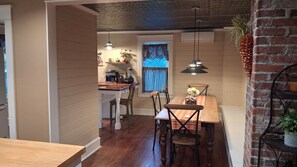 Zona pranzo con tavolo e sedie in legno, parete decorata con mattoni, illuminazione a sospensione e cucina sullo sfondo.