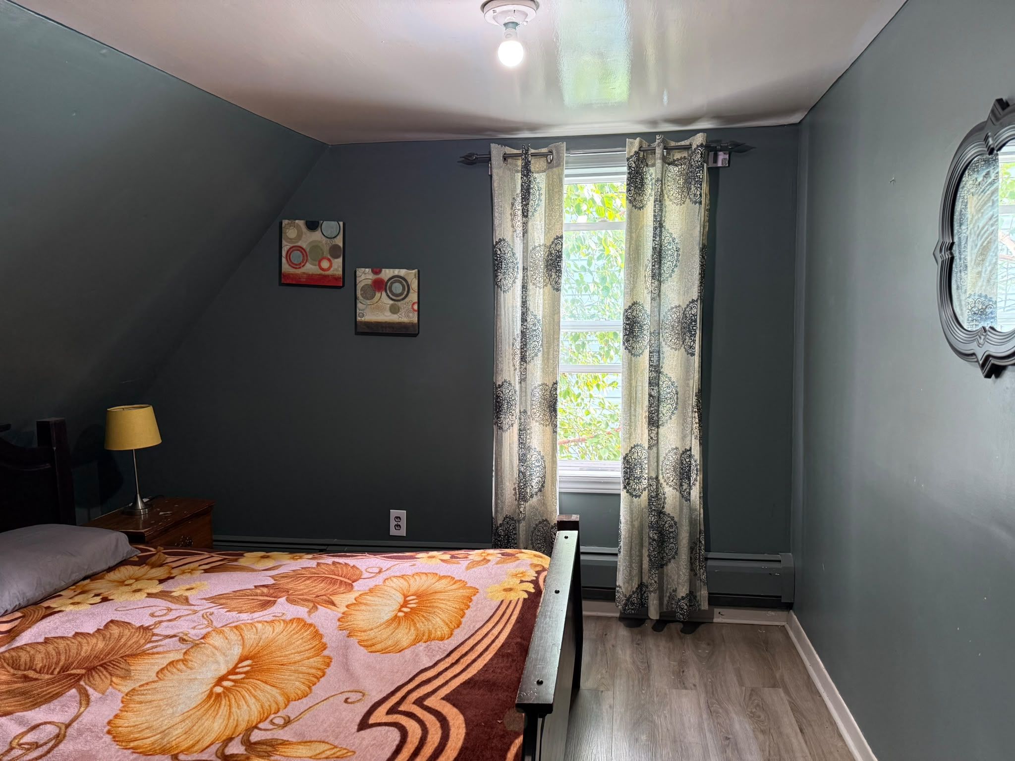 A bedroom with a bed, a nightstand, a lamp, a window with curtains, and two wall art pieces.