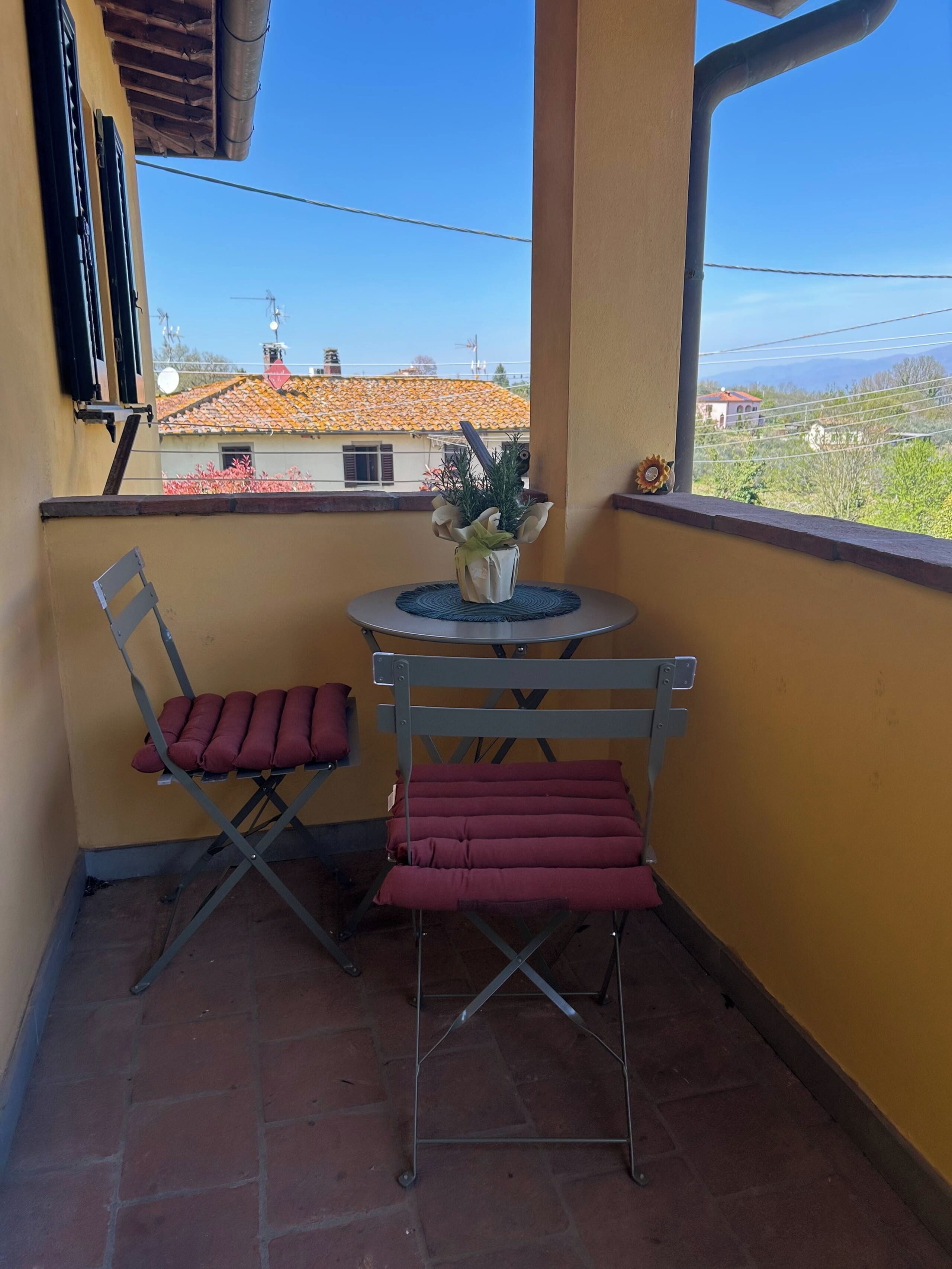 Un balcone con un tavolo, due sedie, una pianta in vaso e vista su edifici e montagne.
