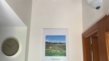 Une pièce avec une poutre en bois au plafond, un tableau encadré représentant la Toscane, une horloge et une étagère sur laquelle se trouvent une bouteille et une bougie.