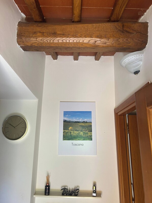 A room with a wooden ceiling beam, a framed picture of Tuscany, a clock, and a shelf with a bottle and a candle.
