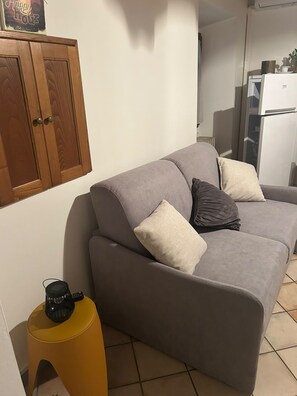 Un divano grigio con due cuscini, uno sgabello giallo e un armadietto in legno con un cartello sopra.