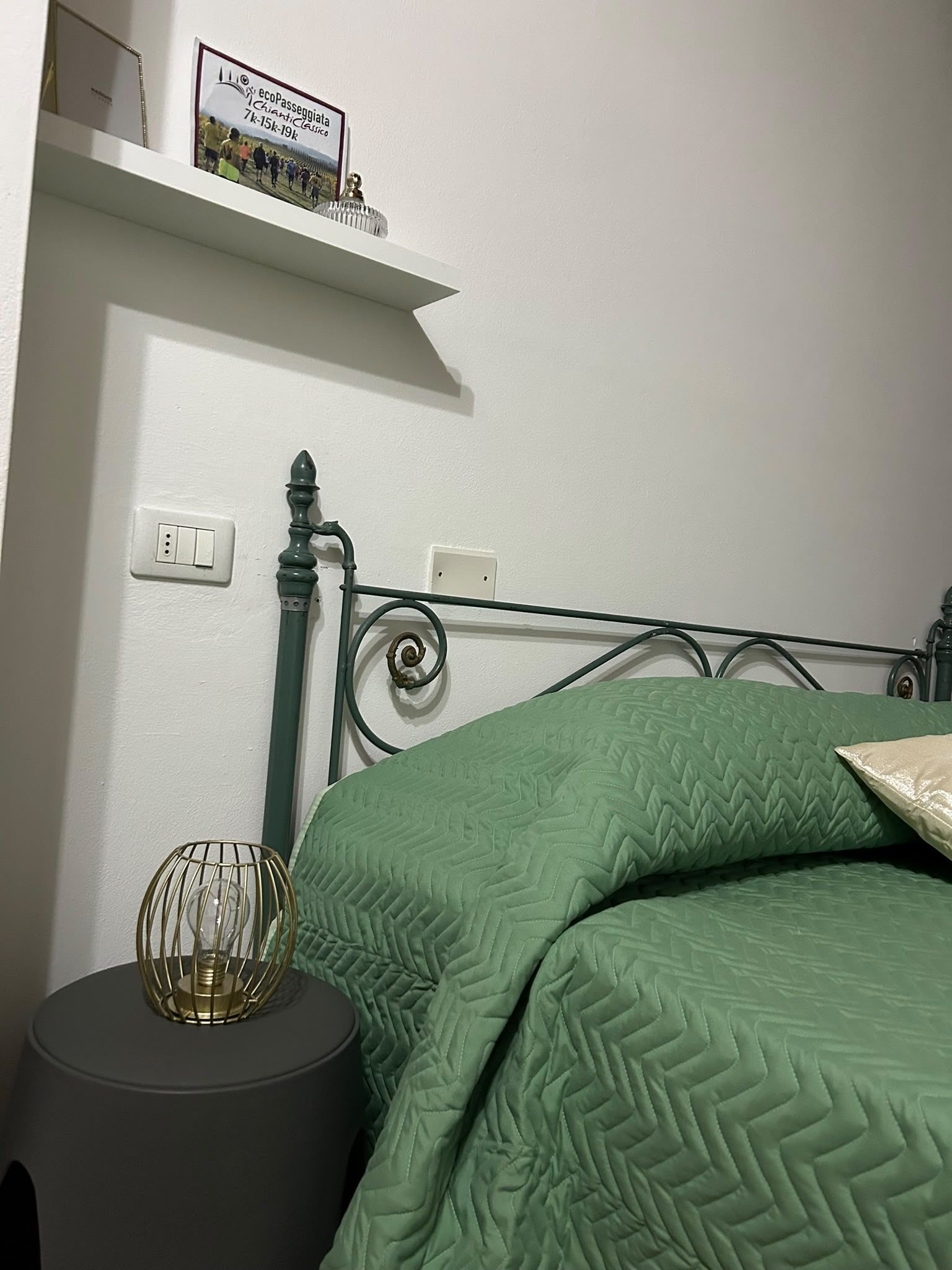 Une chambre à coucher avec un lit en métal, une literie verte, une lampe de chevet et une petite étagère sur laquelle est posé un panneau.