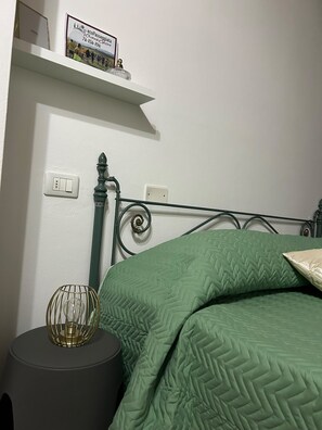 A bedroom with a metal bed frame, green bedding, a bedside lamp, and a small shelf with a sign.