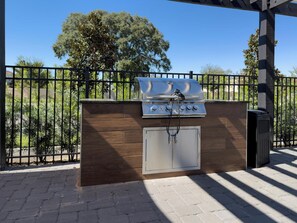 Een buitenbarbecue met een houten kast en een metalen afdekking, op de achtergrond een metalen hek en bomen.