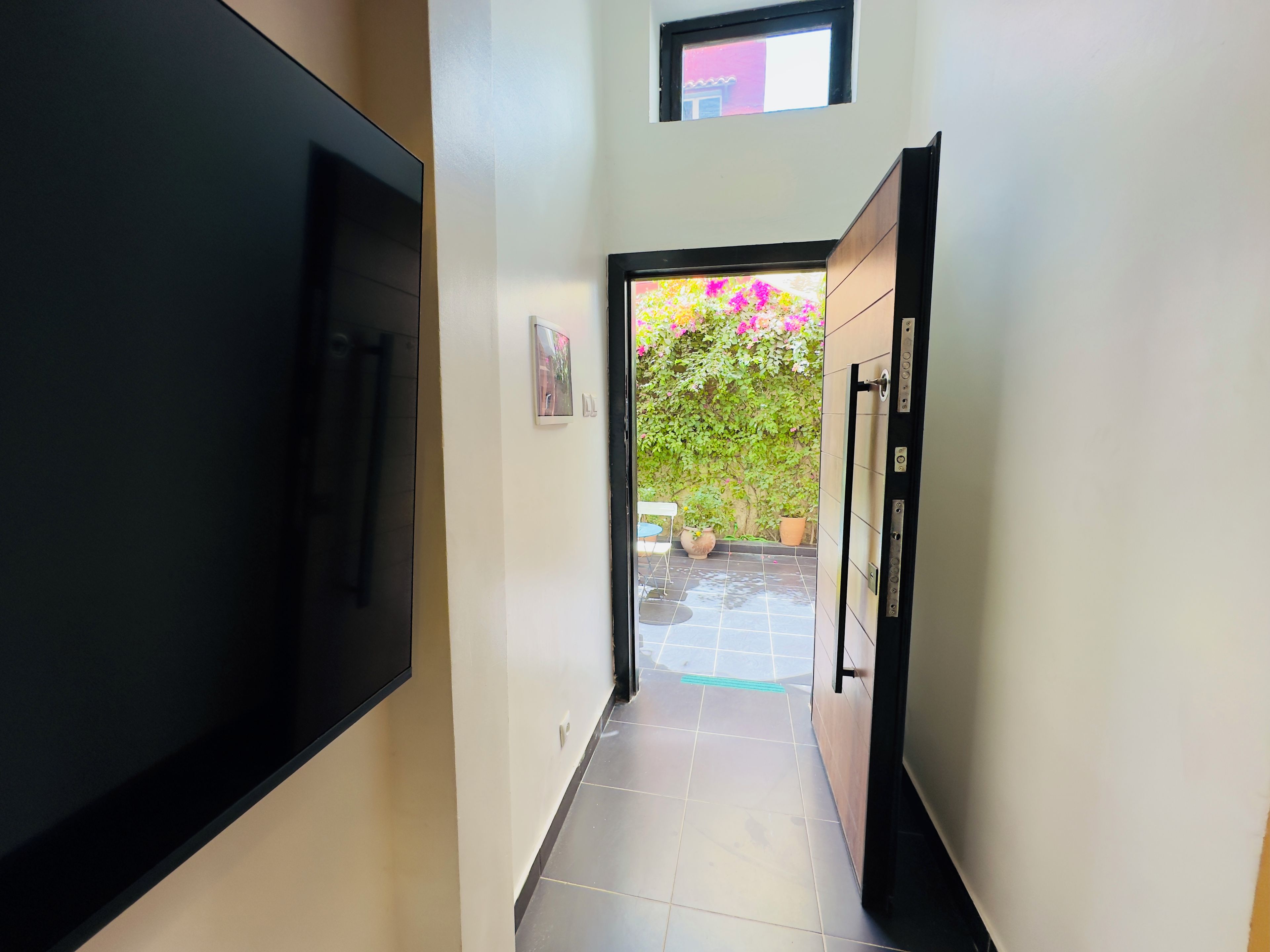 A hallway with a flat-screen TV mounted on the left wall, a door leading to an outdoor area with potted plants, and a tiled floor.