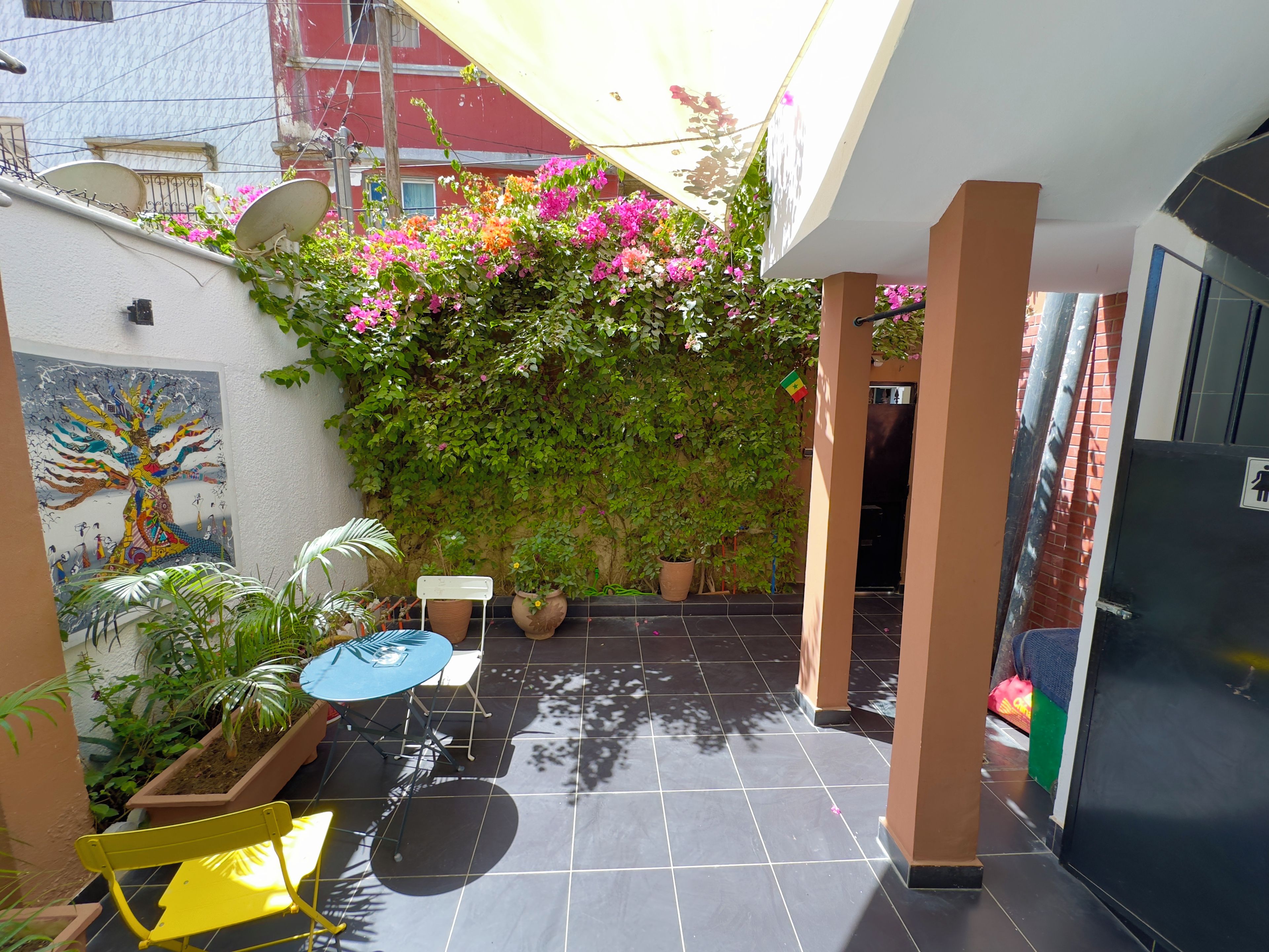 Un espacio exterior techado con suelo de baldosas, una mesa con sillas, plantas en macetas y una pared cubierta de plantas trepadoras.
