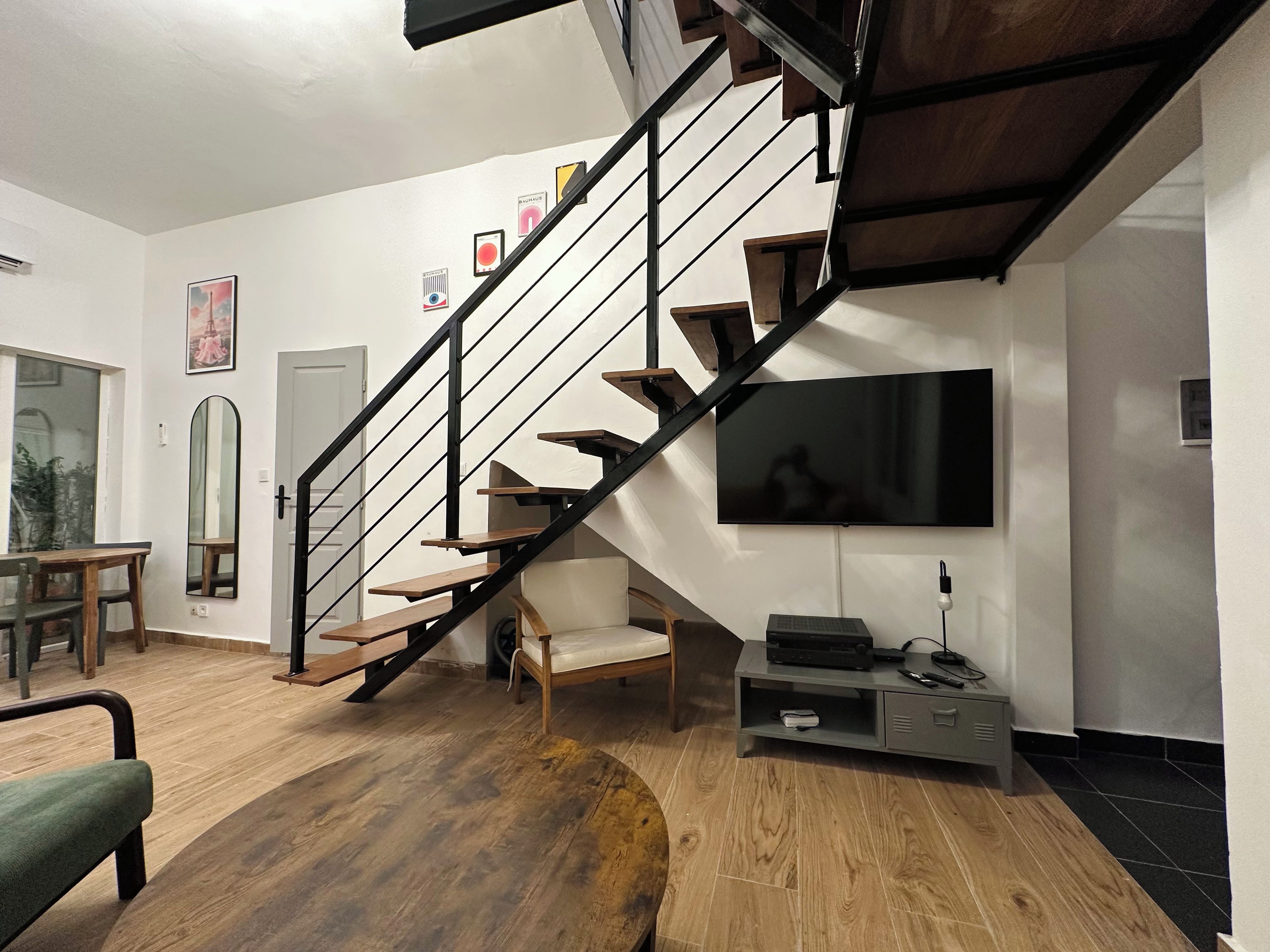 Una sala de estar moderna con piso de madera, una escalera con barandas negras, un televisor de pantalla plana en la pared y una zona de estar con un sofá verde y una mesa de centro de madera.