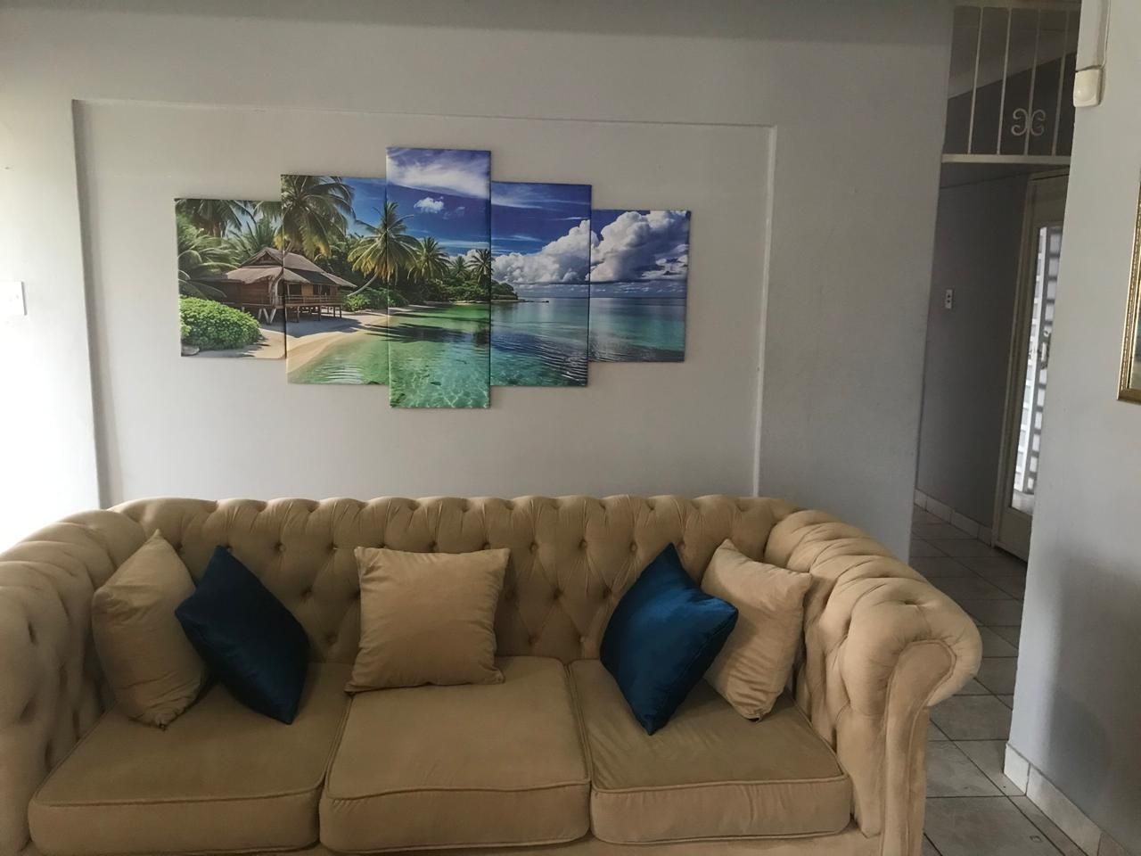 A beige sofa with blue cushions in a room with a large tropical beach painting on the wall.
