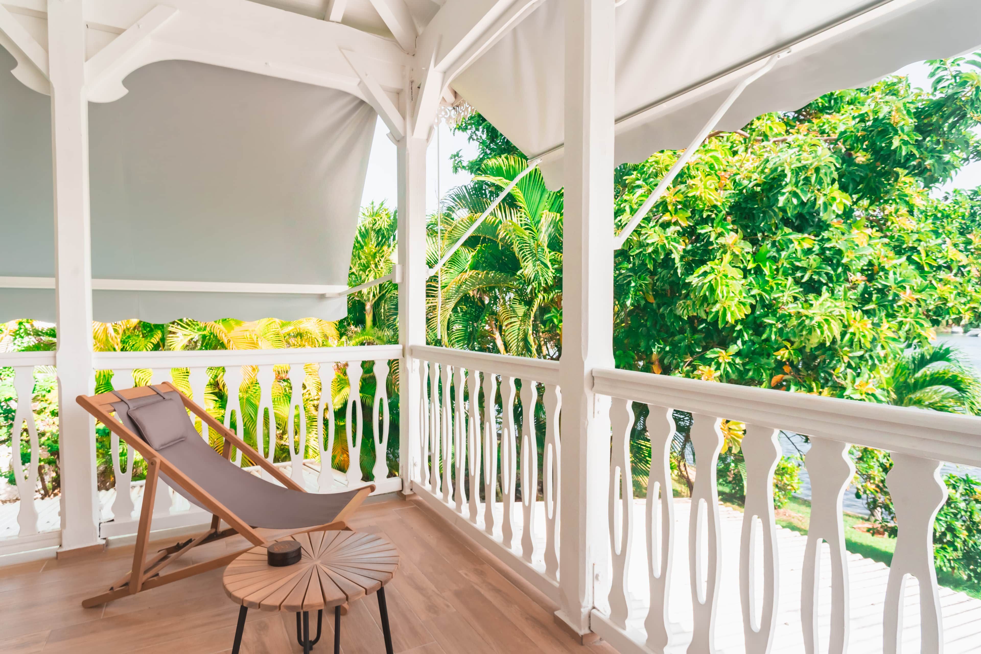 A covered balcony with a wooden chair and a small table, overlooking lush greenery and a body of water.