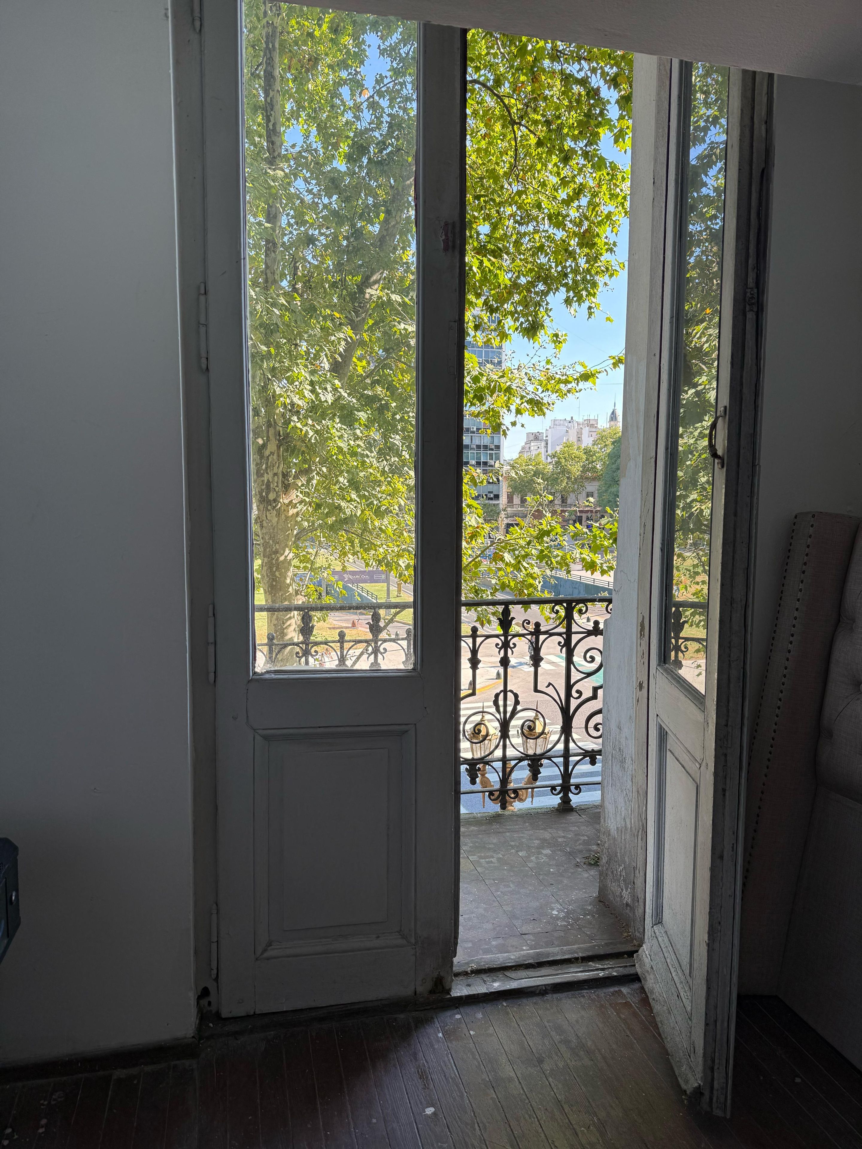 An open door leading to a balcony with a wrought iron railing and a view of trees and buildings.