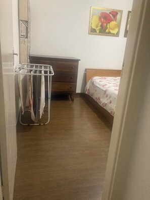 Een slaapkamer met een houten dressoir, een bed met een bloemenprint beddegoed, een aan de muur hangend fotolijstje en een rek met kleding.