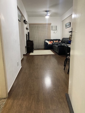 A living room with a dark wooden floor, a black leather sofa, a small table, and a ceiling fan. - Apartment in Guarujá - Enseada Beach - Excellent (Guarujá)
