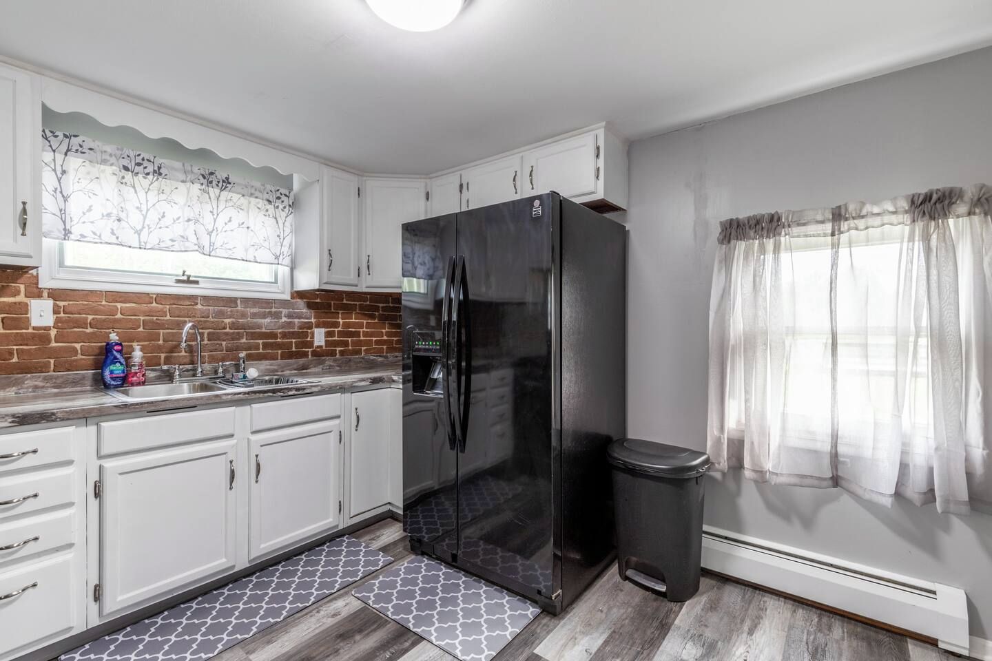 A kitchen with white cabinets, a black refrigerator, a sink, and a window with sheer curtains.