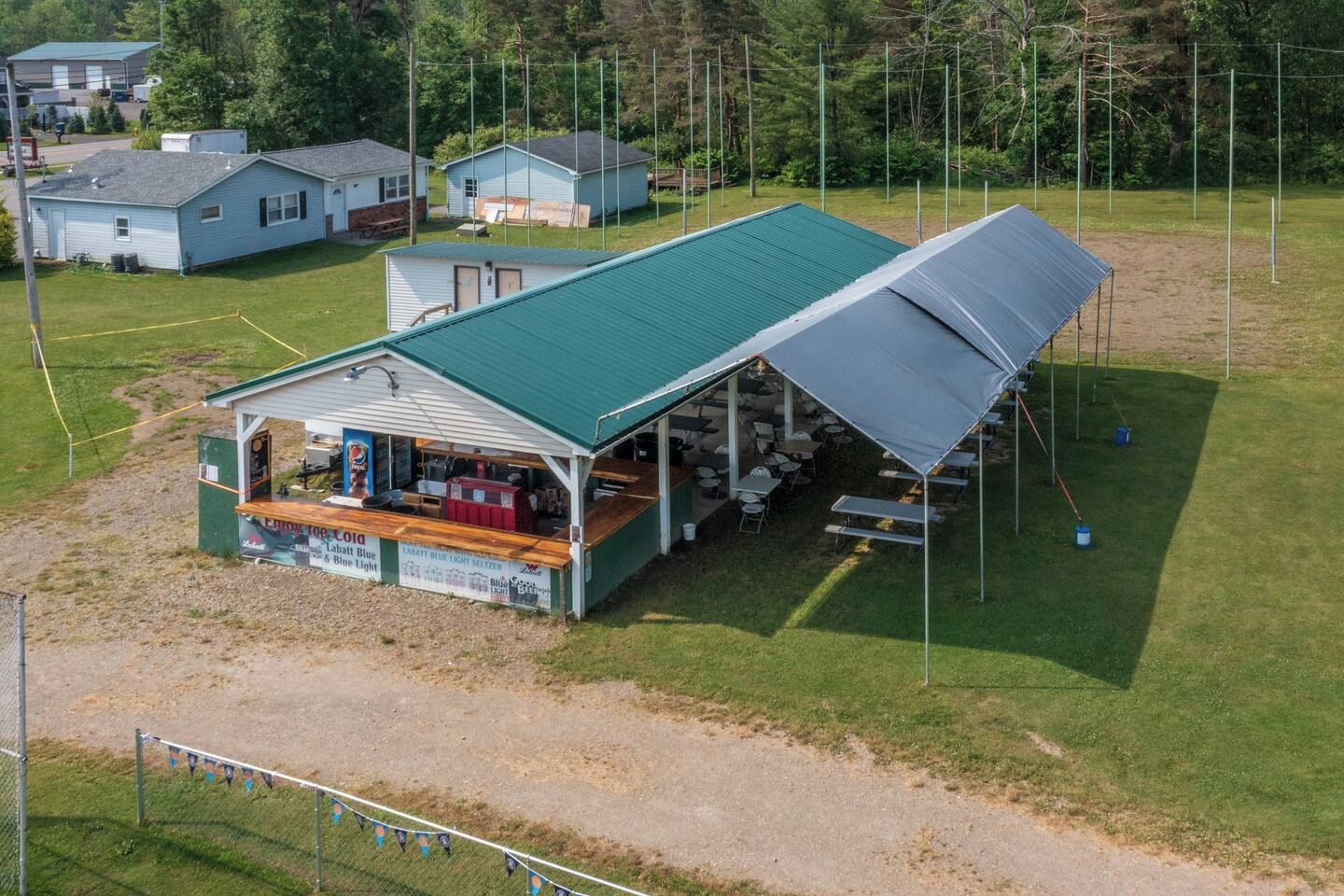 Un petit pavillon extérieur doté d’un toit végétalisé, entouré d’un champ herbeux et bordé de quelques bâtiments en arrière-plan.
