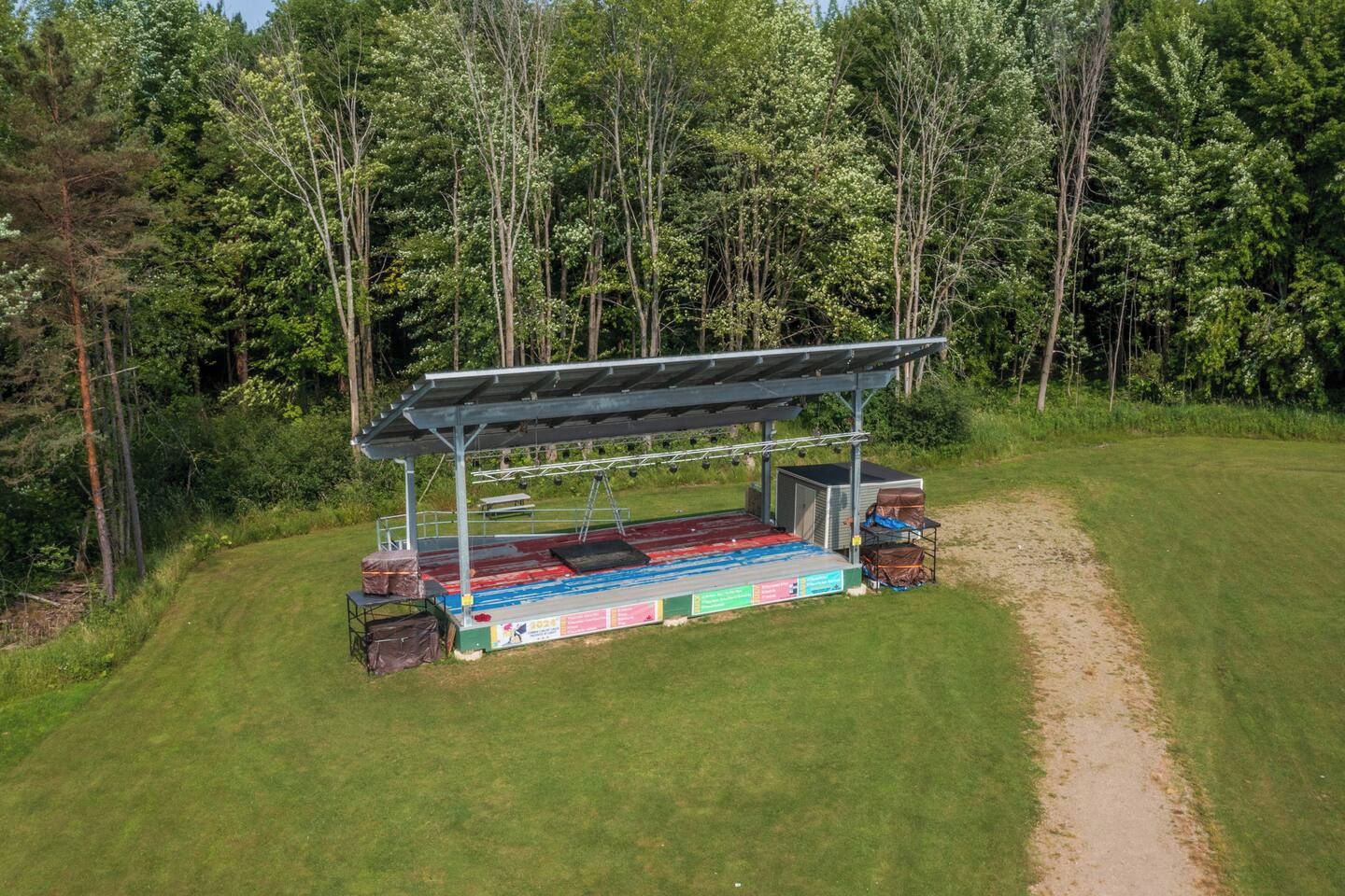 A covered outdoor stage with a flat platform, surrounded by a grassy area and dense trees.