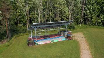 A covered outdoor stage with a flat platform, surrounded by a grassy area and dense trees.