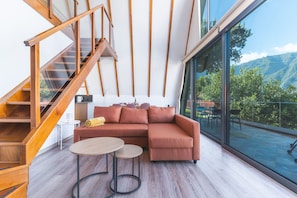 A modern living room with a wooden staircase, a large window with a mountain view, a brown sectional sofa, and two small round tables. - Star Lodge by Atlantic Holiday (São Vicente)