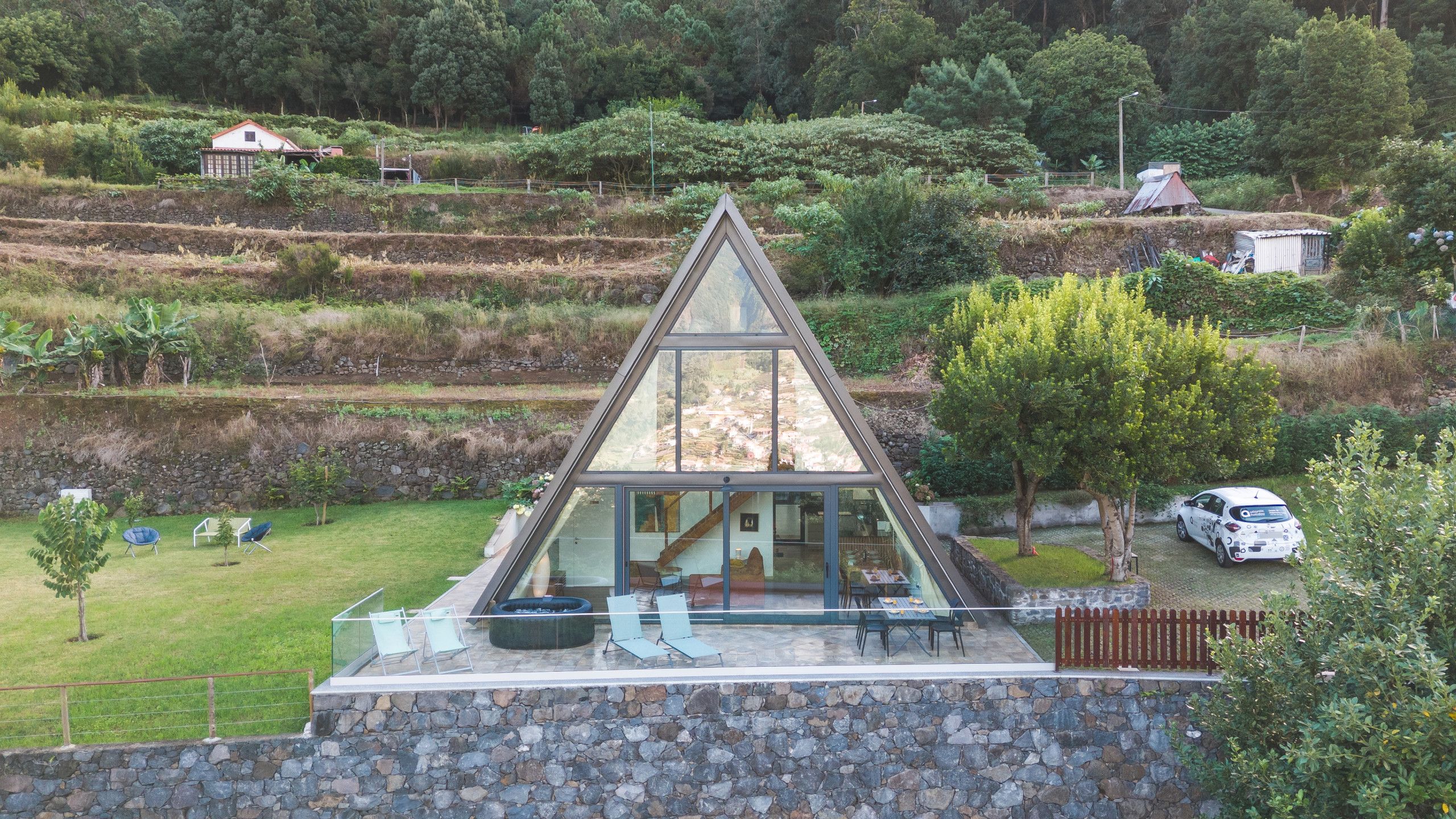 Une maison moderne dotée d’une façade triangulaire en verre, d’un mur en pierre et d’un petit jardin agrémenté d’un espace salon extérieur.